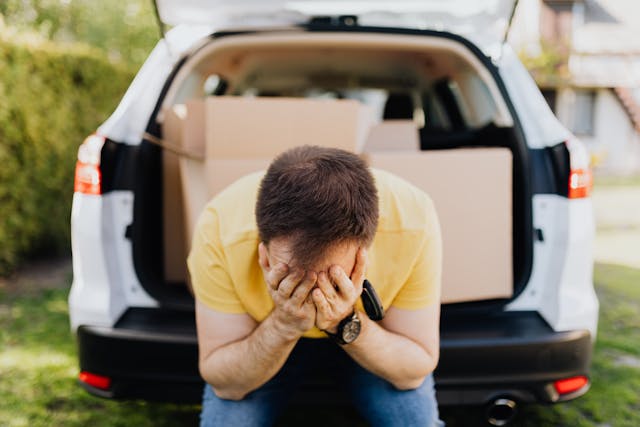 Overwhelmed by Life's Burdens: A Moment of Financial or Emotional Struggle Stressed man sitting on the edge of a car trunk filled with cardboard boxes, covering his face with his hands in frustration or despair.