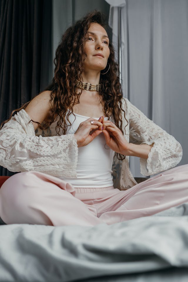 Woman Practicing Mudra Meditation in a Calm Space A woman with long curly hair sits cross-legged on a bed, forming a mudra gesture with her hands during a meditation session.