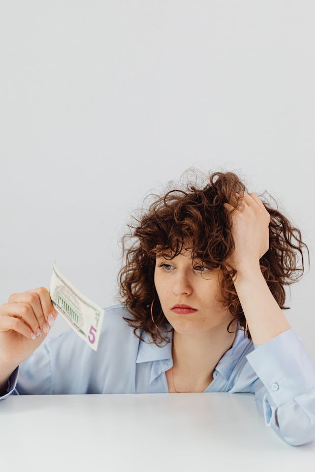 Living paycheck to paycheck Woman looking frustrated while holding a five-dollar bill against a blank background symbolizing feeling trapped in scarcity and the call to wake up from the matrix.