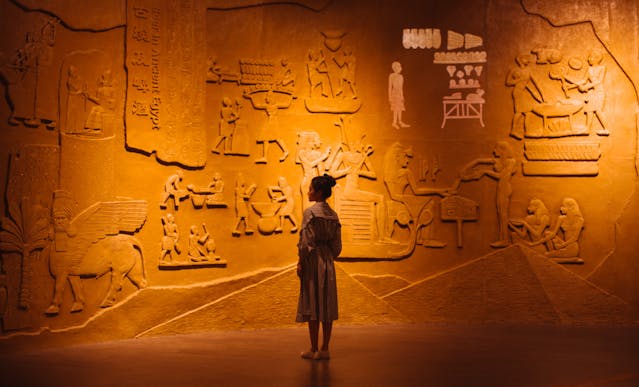 Girl Observing Ancient Wall Carvings in a Golden-Lit Museum A young girl stands alone in front of an illuminated wall covered in ancient carvings and symbols in a museum exhibit.
