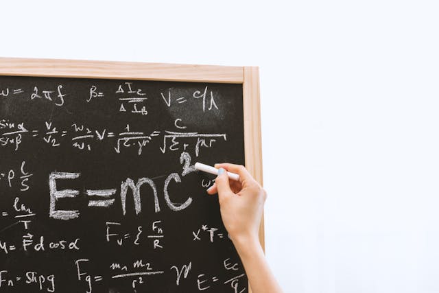 Hand writing E=mc² on a chalkboard surrounded by physics equations in white chalk