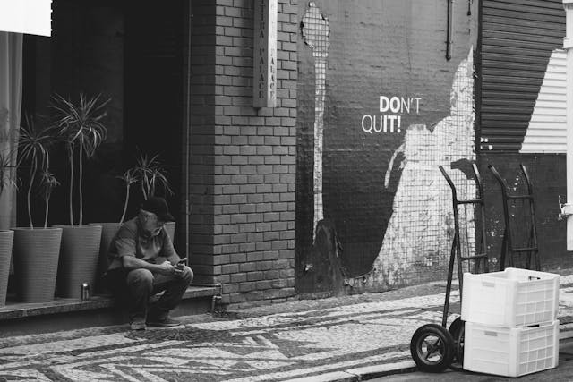 Black and white photo of a man sitting on a sidewalk near street art with the words “Don’t Quit” painted on a wall