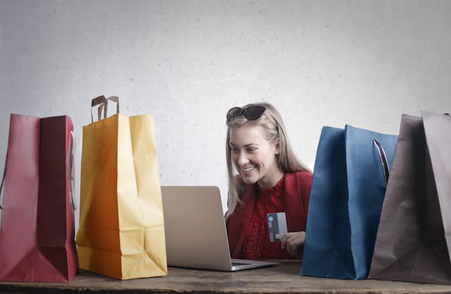 Spend With Purpose, Not Pressure Smiling woman shopping online with bags and a credit card—planning smarter finances to escape the 9 5 rat race.