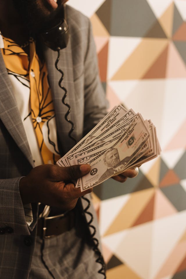 Man in a patterned shirt and gray suit holding a fan of U.S. dollar bills while speaking on a vintage phone, against a geometric wall
