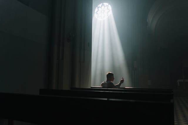Man praying alone in a dim church as a sunbeam pours through a rose window, reflecting on ask believe receive scripture.