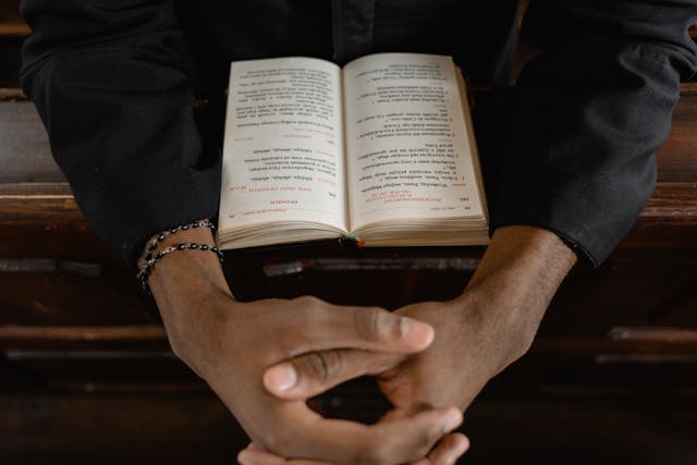 Clasped hands with a rosary resting on an open Bible at a pew, focusing on ask believe receive scripture.