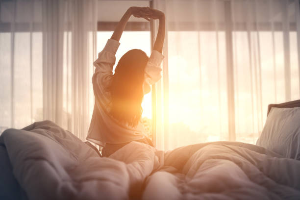 Woman stretching on her bed in the early morning sunlight shining through sheer curtains.