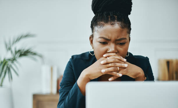 Focused woman sitting at a laptop with hands clasped in thought, reflecting on challenges before shifting her mindset using abundance mindset affirmations.