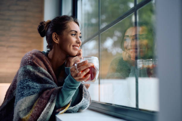 Woman smiling while looking out the window with a warm drink, showing peaceful signs manifestation is working.