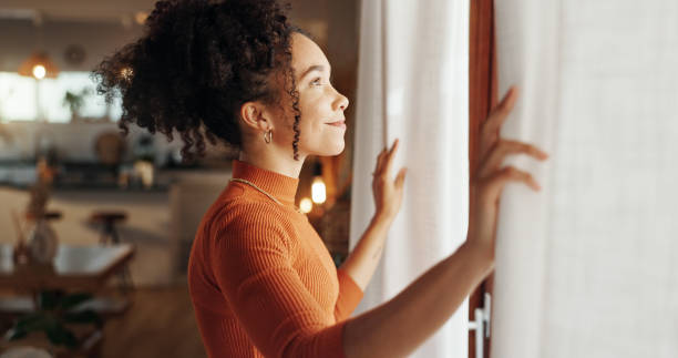 Smiling woman in an orange sweater opening white curtains to let in morning light, representing positive identity shifts and new beginnings.