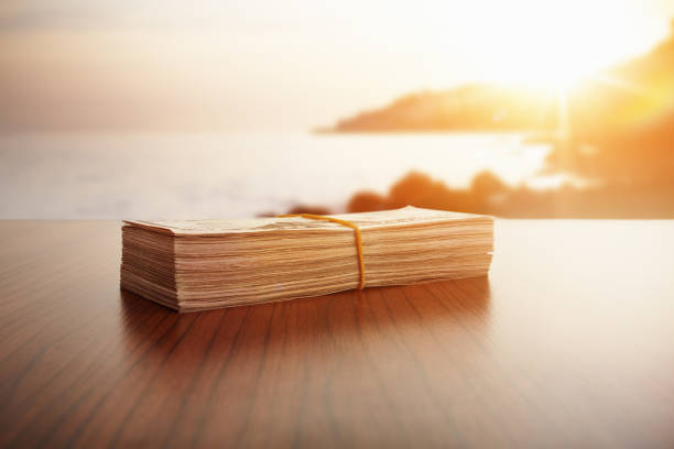 Stack of cash sitting on a wooden table with a glowing sunset over the ocean in the background.