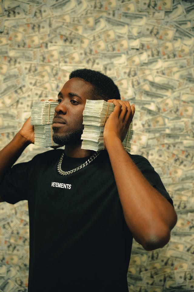 A man holds stacks of cash beside his ears, standing before a wall covered in dollar bills, symbolizing wealth attraction through money magnet affirmations.