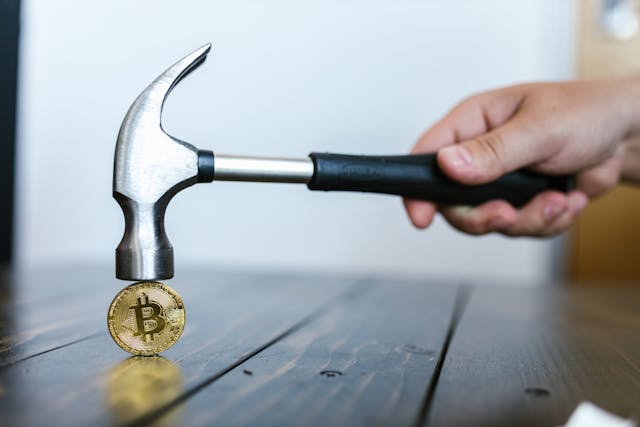 A hand holds a hammer above a gold Bitcoin coin on a wooden surface, symbolizing breaking financial fears and reprogramming wealth beliefs through money magnet affirmations.