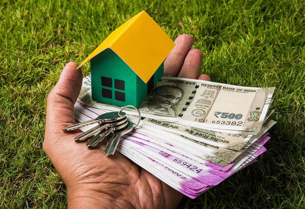Manifesting a Home with a Money Blessing Prayer A hand held over green grass carrying a stack of Indian Rupee notes, a small green toy house with a yellow roof, and a set of silver keys.