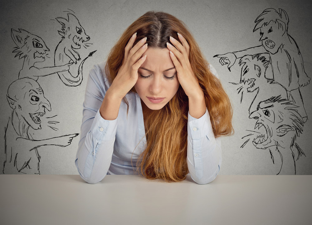 Stressed woman holding her head while sketch-like figures around her shout and criticize, representing negative self-talk and internal doubt.
