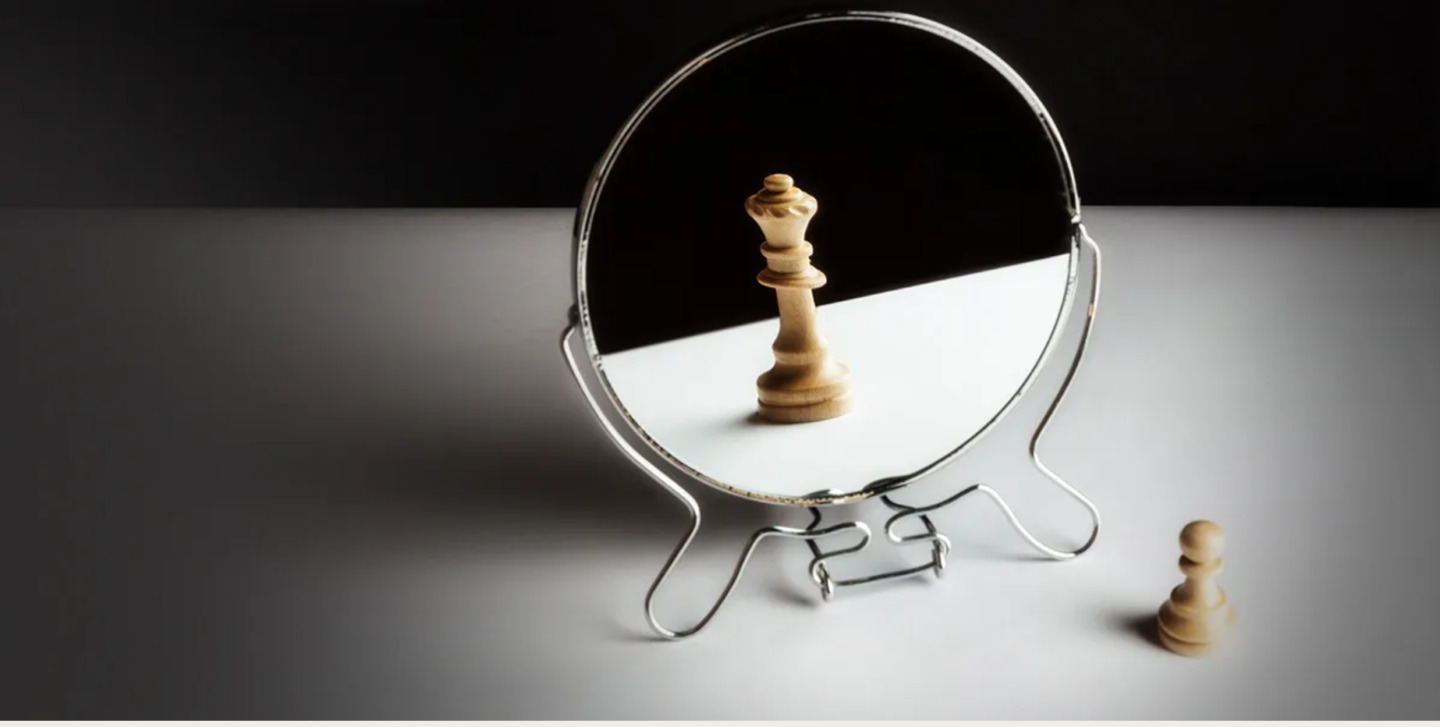 A small chess pawn sits beside a round mirror, where its reflection appears as a queen against a dark, minimalist background.