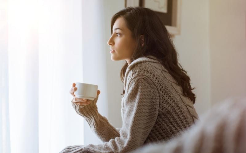 Woman sitting by a window holding a cup of tea and looking outside while reflecting on her thoughts and emotions.
