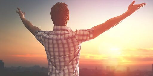 A man seen from behind with his arms raised wide toward a glowing sunset over a city skyline.